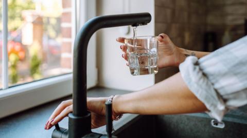 Someone pouring water into a glass from a black tap. You can only see their arms and hands. There is a window in the background.