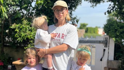 Jenna Weber is standing in her garden, next to a road. She is holding her youngest daughter while her other two daughters stand next to her.