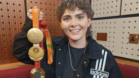 A woman with curly brown hair holds up three gold medals. She is wearing a a black t-shirt, a black Adidas jacket, a gold chain around her neck.