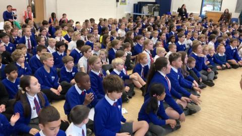 A group of school children wearing blue and white school uniform. They are sat on the floor of a school hall.