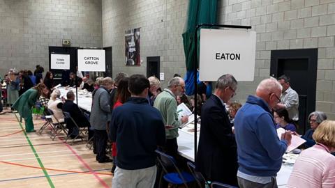 People milling around a line of trestle tables at an election count in a sports hall