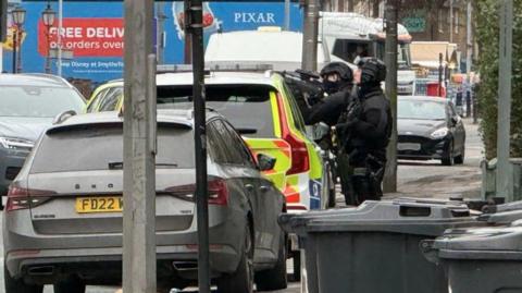 Two armed police in black outfits and helmets are standing on a street in front of a police car. One is looking up and the other is aiming a gun.