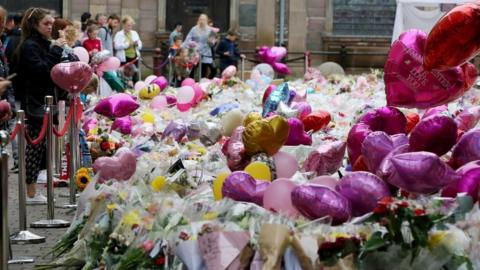 People stand at a memorial in central Manchester following the Arena bombing. There are balloons and flowers lying on the ground.