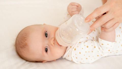 Baby wearing a white and yellow babygro lies on its side with a bottle of infant formula to its lips, held there by an adult's hand