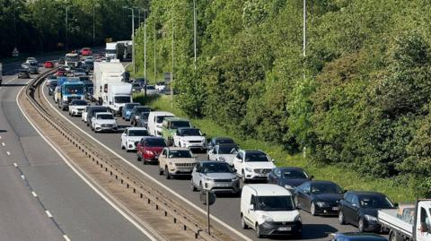 A queue of traffic along both lanes of a dual carriageway heading from the top left to the bottom right of the picture. The traffic includes lorries, cars and vans. There is a low barrier in the centre of the road and some vehicles are visible heading in the opposite direction in the top right. There are trees to the right of the photo and lamp poles.