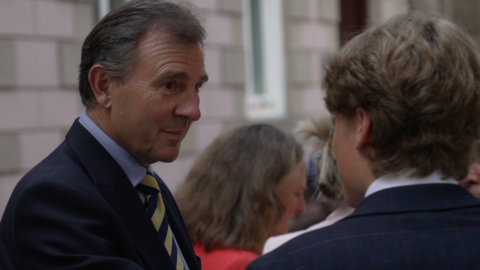A person in a dark suit and striped tie speaks with another person in a suit at an indoor event, with several people blurred in the background.