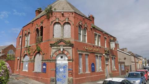 A derelict, two-storey redbrick pub stands on a corner of a terraced street. The windows are either missing or boarded up and plants are growing from the brickwork. Blue signs, and some blue graffiti can be seen. The name "Queen's Arms" can be seen on two sides of the building in gold lettering, some of which is broken.