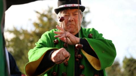 David Jakins is dressed in green and adorned with conkers. He is focusing on a game of conkers he is currently playing. He is wearing a black hat, and there are trees behind him. 