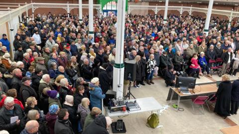 A view from above the crowd, showing people standing shoulder to shoulder across the hall.  The people are in their coats and jackets, facing the speakers who are stood behind a table.