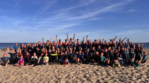 A large group of children and adult volunteers with their arms in the air. They're all on the sand at Branksome Chine beach with the sea behind them. They're wearing black wetsuits and orange swimcaps.