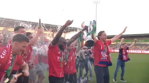 A football team wearing medals, dressed in a red strip, lifting a silver trophy together.