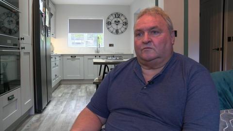 A man in his 60s, wearing a navy polo shirt, sits on a chair in a kitchen while being interviewed by the BBC.