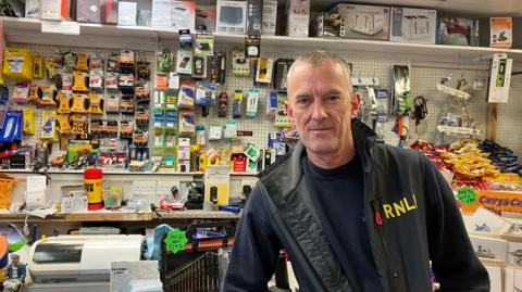 A man in a shop. He is wearing an RNLI jacket. Behind him are crisps and batteries and different appliances on shelves. There is a till behind him. 
