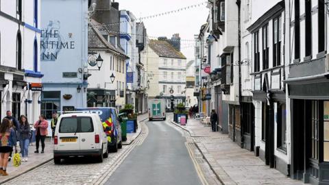 A picture of a long street in Plymouth. There is shops pictured either side of the road with a number of vehicles pictured in the image. There is also people walking on the footpath.