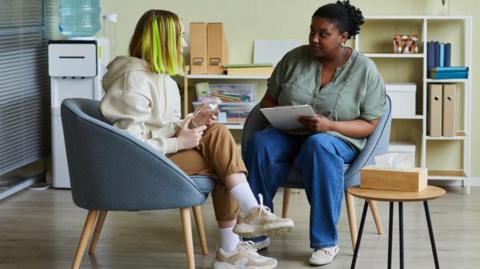 A teenage girl sits in a chair and talks to a social worker, who is holding papers and listening to her. They are in an office and there are shelves behind them with a stand for drinking water.