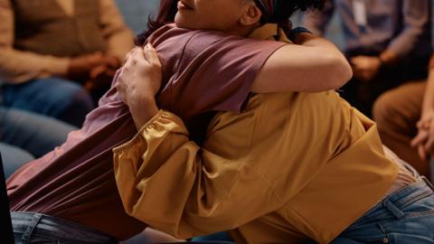 Two people sat on chairs in a group lean into each other for a hug. The person on the left is wearing a maroon top while the person on the right is wearing a yellow blouse with puffed sleeves. 