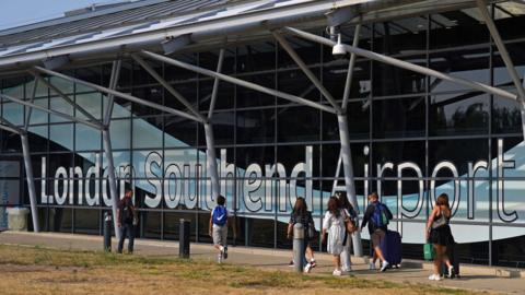 Passengers outside the terminal building at Southend Airport. the building is a mostly glass structure. People wear summer clothes and it is sunny.