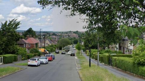 A road with houses on both sides of the street and in the distance. Cars are parked on both sides of the road. There are grass verges and trees on both sides of the street.