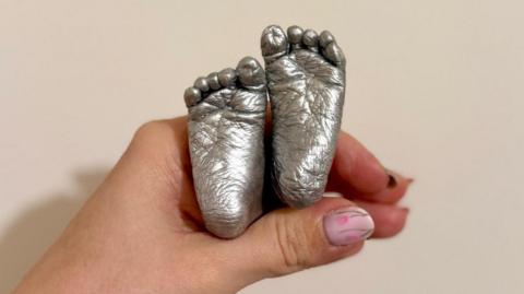 Two small, pewter baby feet are held in a woman's hand against a plain background.