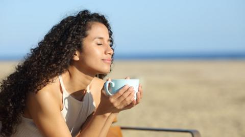 A woman holds a cup of tea on a beach, her eyes are closed and the sun is on her face