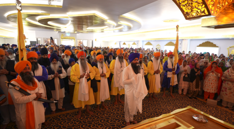 Dozens of male Sikhs in traditional dress gather inside the temple with a large crowd of men and woman behind them. The men have yellow, red, blue and orange turbans on, some holding flags. There is a gold design on the ceiling and walls.