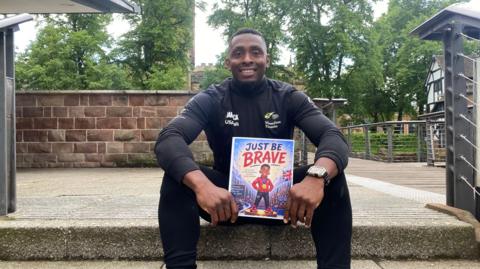 Joel Fearon, who is dressed in sports gear is sat on a stone step, with a wall and trees behind him. He is smiling at the camera and holding up his book, which is called Just Be Brave.