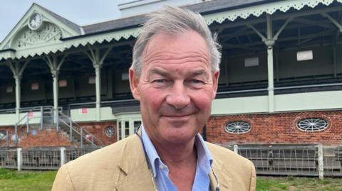 Rupert Lowe, a man standing outside. He is looking directly at the camera and smiling. He is wearing a tan linen suit jacket with a pale blue shirt.