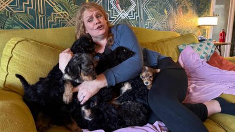 A blonde woman cradles numerous black and brown puppies on an yellow sofa.
