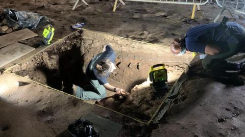 Archaeologists at a site are digging in a rectangular hole in the ground surrounded by metal fences. They are wearing head torches