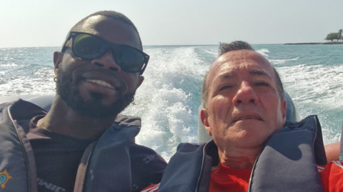 Yostin Mosquera (Left) and Albert Alfonso (right) on a speedboat. They are wearing lifejackets and smiling at the camera. There is clear blue sea behind them with the wake of the waves. In the distance palm trees can be seen on the shore line. 