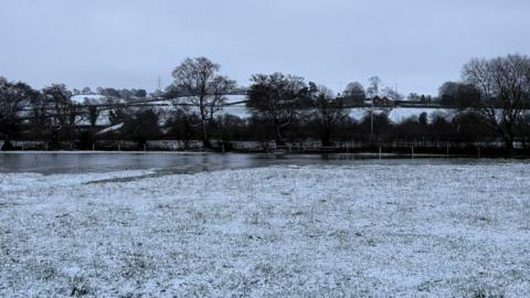 Grass covered in snow, with water, trees and hills in the distance.