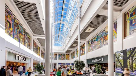 A central square in a shopping centre with a number of people pictured from the back. A Next and Coffee#1 is visible and there are also indoor trees and plants. Around the top of the atrium is a brightly coloured mural. 