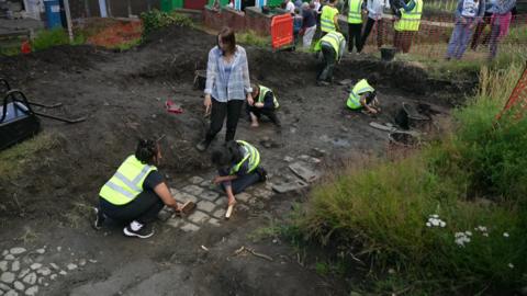 Image shows people in high-visibility tabards digging into the earth, uncovering what look like sections of cobbled streets below the soil