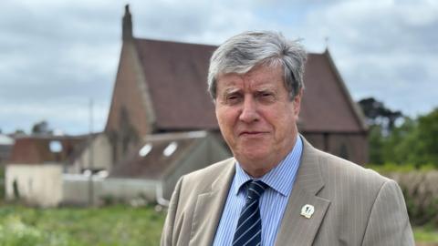 Kevin Lewis, standing in the field that could be developed. The parish hall is visible in the background. Kevin is wearing a brown suit jacket, blue shirt and navy tie. 