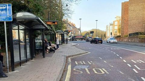 The bus stops on Milburngate. The shelters have been built in a row meaning people would have to step out to see arriving buses. A small group of people wait at the middle bus stop, including a woman with a pram. There are a few vehicles at a junction further up the road, which is in part a reddish brown colour to signify bus stops and a bus lane.