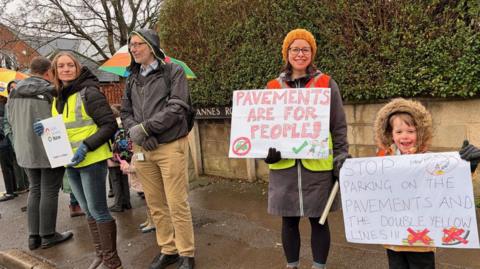 A group of people stand on a pavement holding signs. Becky Carlyle stands to the right with her son. Her sign reads: "Pavements are for people." Her son's sign says: "Stop parking on the pavements and the double yellow lines."