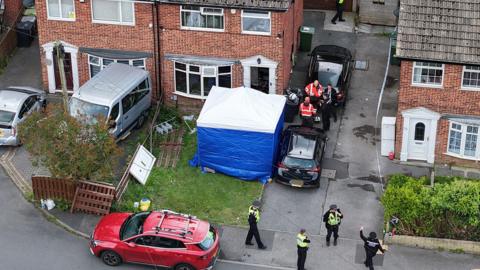 Police gather in front of a row of semi-detached properties. One house has a blue police tent in the front garden and has the door missing. 