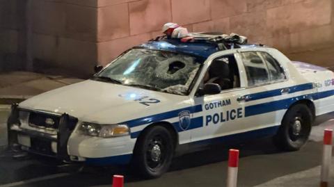 A blue and white liveried police car emblazoned with the sign of Gotham Police. the front windscreen is shattered with a hole in the glass.