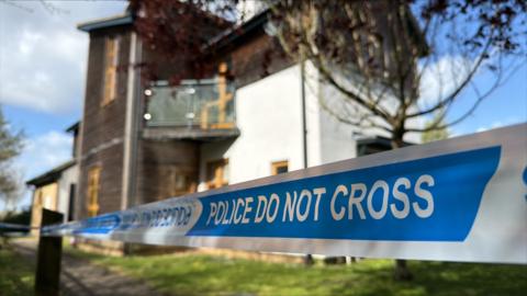 Blue and white tape stating POLICE DO NOT CROSS stretches across the front of the image. Behind is a blurred background of a white house with dark wooden cladding.