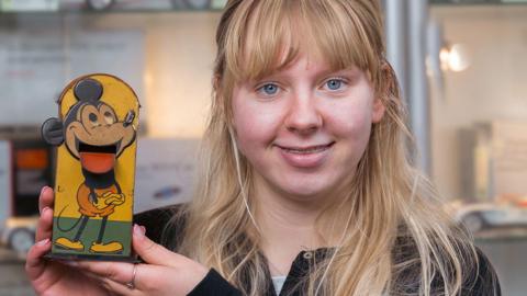 A woman smiles while holding a vintage money box painted with a cartoon mouse, standing in front of a display case filled with model cars and other collectibles.