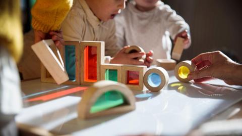 Three toddlers play with coloured shapes around a table at a nursery.