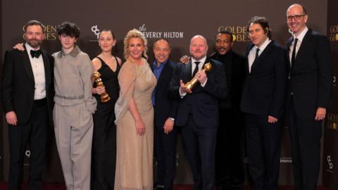 A group of eight individuals standing together on a red carpet. The backdrop behind them features multiple logos, including “Golden Globes”, “CBS,” “Paramount+”, and “The Beverly Hilton”.
Several people in the group are holding Golden Globe trophies. The individuals are dressed in formal attire, including tuxedos, suits, and elegant gowns.
The group is posed closely together, with some placing their arms around each other.
