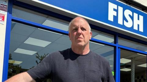 A man wearing a dark t-shirt stands outside his chip shop.