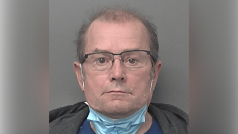 A police custody photo of Darren Richardson. He has short, sparse brown hair and is wearing black-rimmed glasses and a face mask under his chin, with the straps around his ears. He is also wearing a blue T-shirt and a grey hoodie. The wall behind him is grey.