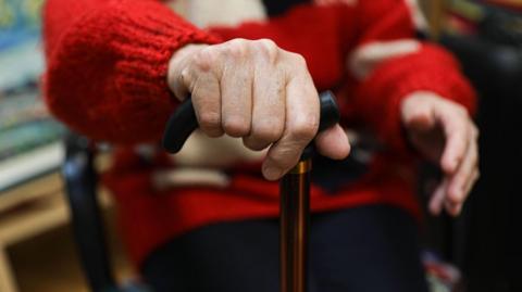 A hand is holding on to the black handle of a walking stick. The person is wearing a red knitted jumper. 