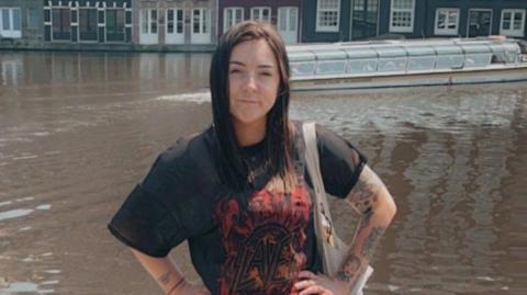 A woman with long black hair and wearing a black t-shirt with her hands on her hips stands in front of a canal.