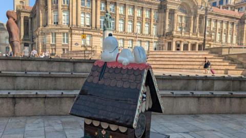 A sculpture of Snoopy the cartoon dog lying on a small wooden house, which is placed in front of Birmingham's Town Hall.