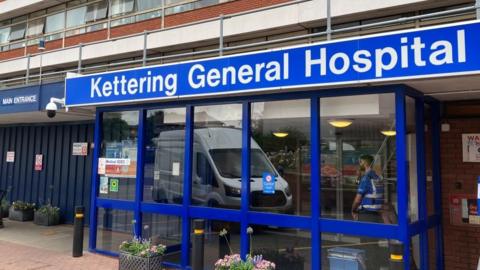 The entrance to Kettering General Hospital, showing blue-framed windows in the lobby and a Kettering General Hospital sign above. A sign saying "main entrance" is visible to the left, above a boarded-up section of wall. A man in a white and blue hi-vis vest and a white van are reflected in the glass of the entrance lobby.