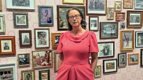 A woman in a pink dress stands against a backdrop of small photographs on a pink and white wall
