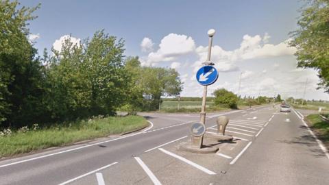 The junction of a main raid with two side roads, surrounded by trees and green trees. The grey asphalt is split by a crossing point with blue signs telling motorists to keep left. White road markings denote the turn-offs.